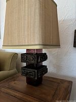 Full view of the lamp showing the reddish glass base, black metal brutalist geometric accents, and the cream rectangular shade on top.
