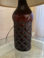 Close-up photo showing the brown tinted glass body of the table lamp with decorative black metal overlay in a quatrefoil pattern. The photo focuses on the lamp's main body design.