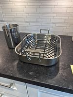 Photo of All-Clad roasting pan with removable black roasting rack inside, and separate stainless steel utensil holder placed beside it on a kitchen counter.