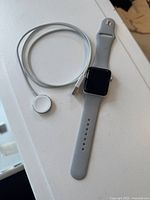 Apple Watch laid on a white surface with gray sport band and magnetic charging cable placed beside it.