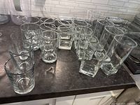 Multiple clear glass drinkware including lowball, highball, and square base glasses arranged on countertop.