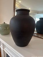 Side angle view of black terracotta vase, showing traditional shape and matte finish, placed on a white surface with mirror and green ceramic jar in background.
