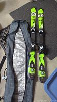 Top-down photo showing pair of green and black Corbetts skis with bindings beside open plaid travel bag on floor.