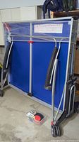 Folded ping pong table in indoor storage position with paddles hanging and plastic container of paddles and balls at base.
