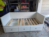 White wooden day bed frame with panel headboard and slatted mattress support, featuring three pull-out drawers along the front.