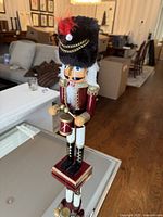 Front angle of wooden nutcracker drummer figurine showing detailed red and gold uniform, black fur hat with chains and red feather, holding drumsticks poised on drum.