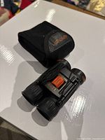 Compact Night Owl binoculars with black casing and orange accents, magnification marked as 8x21, lying on a white surface next to soft carrying pouch.