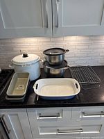 Photo showing all items on black countertop against white tile backsplash, including large white enamel soup pot, stacked stainless steel bowls, baking racks, and baking tins with label sizes.