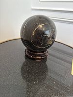 Front view of the black marble sphere with visible natural white veining on its polished surface. Sphere placed on the detailed carved wooden stand.