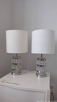 Front view of the pair of lamps with clear and chrome bases with white linen shades sitting on white table.
