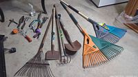 Full view of assorted garden tools laid on concrete floor including rakes, shovels, cultivators, pruners, and hand tools.