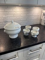 White soup tureen with lid and four matching soup bowls displayed on kitchen counter.