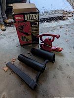 Photo showing Toro Ultra electric blower vac, three black attachment tubes, and original box outdoors on concrete floor with some snow in background.