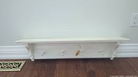 Front view of white wooden wall shelf with four pegs, showing distressed paint and decorative bracket supports.