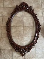 Full view of the ornate oval wooden mirror frame laid flat on tiled floor, showing entire frame and open center.