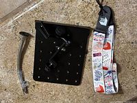 Photo showing black metal mounting plate with adjustable arm, flexible gooseneck clamp, and guitar strap laid out on floor.