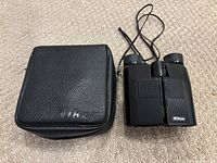 Photo of Nikon binoculars displayed beside their black zippered carrying case on beige carpet.