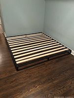 King size platform bed frame constructed with wooden slats on black metal rectangular frame standing on dark hardwood floor in a room with light blue walls.
