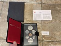 Overview of black leatherette case with red velvet lining containing the coin set, arranged in two plastic trays with a total of 14 coins.
