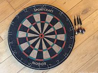 Image of Sportcraft Nodor dartboard showing bullseye, segmented scoring areas and numbering around edge, with four FG Bradley steel tip darts placed beside it on wooden floor.