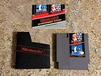 Front side of NES cartridge, manual, and protective black plastic case placed on a countertop showing the game label and manual cover.