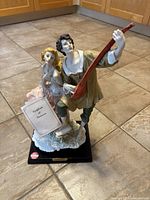 Front view of porcelain musical couple figurine showing male playing stringed instrument and female holding card, on wooden base.