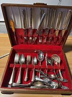 Full view of 1881 Rogers silverware set in an open wooden box with red velvet lining, showing forks, spoons, and knives. Some cutlery pieces are loose at the bottom of the box.