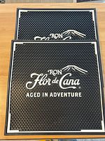 Two black rubber bartender mats shown stacked, featuring 'Ron Flor de Caña Aged in Adventure' logo in white with mountain design, textured surface and white borders.