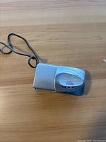 Silver Olympus digital camera lying on wooden surface showing front side including the lens cover and wrist strap.