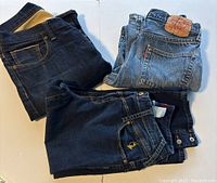 Three pairs of folded jeans: one dark wash, one light blue Levi's with leather patch, and one medium-dark wash with button details.