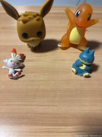 Photo showing four Pokémon figures on a wooden surface: large brown Eevee Funko Pop, orange Charmander, small white and orange Scorbunny, and small blue Totodile.