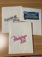 Three press kit covers titled The Flamingo Kid, Unfaithfully Yours, and Moving Violations laid on a wooden surface.