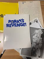 White folder labeled 'PORKY'S REVENGE!' along with typed production notes and several black and white still photographs spread out on a brown surface.