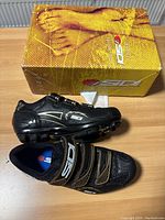 Pair of black Sidi mountain biking shoes with velcro straps, displayed next to branded yellow Sidi box on wooden surface.