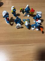 Seven vintage Smurf figurines arranged on a wooden surface showing various poses including holding gifts and flowers.