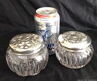 Front view of both dresser jars next to a soda can for size comparison showing crystal bodies and floral embossed sterling silver lids
