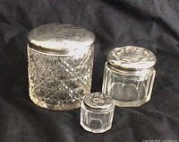 All three cut crystal dresser jars with sterling silver lids of varying sizes shown together on black fabric background; largest jar is heavily cut with floral pattern lid, medium jar hexagonal with engraved initial B lid, smallest jar small and faceted with worn lid and hole near edge.