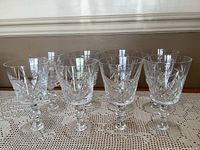 Front view of all 8 crystal glasses arranged in two rows on a crocheted lace tablecloth, showing their pinwheel cut design and thick stems with circular protrusions.