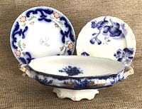 Front view of entire lot showing two decorative plates (one with blue and peach floral pattern, one with blue floral pattern) and large footed oval serving bowl with blue floral design and gold trim on handles and rim, slight wear visible.