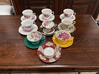 Top view of all 9 vintage floral cups and saucers arranged in 3 rows on wooden table showing color and pattern detail