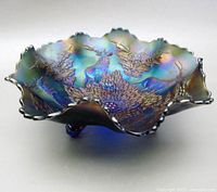 Front and angled side view of iridescent blue carnival glass bowl showing stag and holly motifs in high relief. Scalloped rim and iridescence visible.