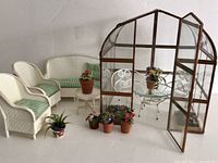 Photo showing two white wicker armchairs with green striped cushions, loveseat, small round side table with potted flowers, six potted miniature plants, and a greenhouse with glass door open.