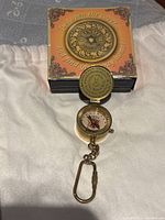 Compass open on cloth surface showing the compass dial with star and directional markings. The box behind features an inscription.