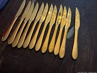 Set of twelve gold-tone steak knives laid out horizontally on a dark wood surface; varying in blade shape but uniform in gold finish with serrated edges.