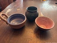 Three signed pottery pieces displayed on a wooden table: a handled cup with blue glaze, a dark glossy vase, and a reddish-brown bowl.