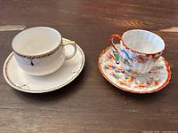 Two vintage Japanese tea cups with saucers displayed on a wooden surface. One white with gold and blue floral accents, the other colorful with hand painted orange scalloped edges and traditional Japanese motifs.