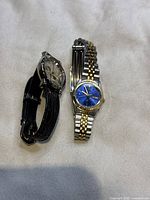 Photo showing two watches side by side: one with a black leather strap and cherub face, the other a two-tone metal bracelet watch with blue dial.