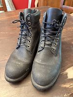 Front view of the pair of black Timberland leather boots on a wooden table, showing scuffed but intact leather and laces tied.