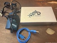 Photo of Phantom Drives GreenDrive 1TB external hard drive, power adapter, and blue USB cable laid on wooden surface.