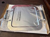 Square stainless steel platter in original box and plastic protective covering, showing front view with handles visible.